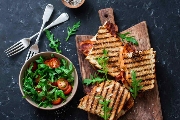 Grilled bacon, mozzarella sandwiches on wooden cutting boards and arugula, cherry tomato salad on dark background, top view.Delicious breakfast or snack, flat lay